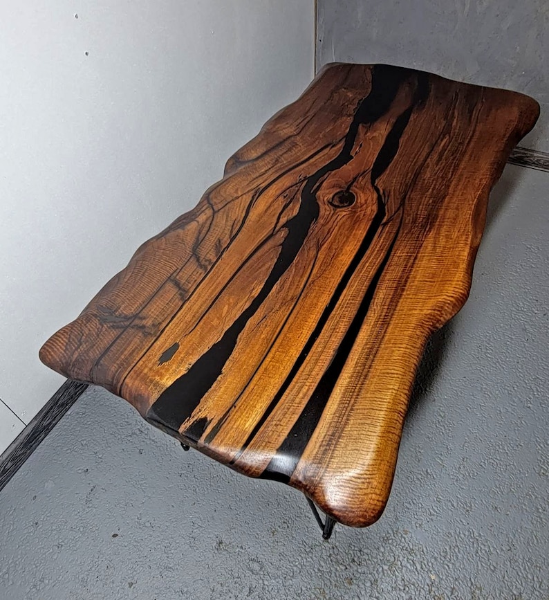 May include: A live edge coffee table with a natural wood top and black epoxy resin filling the cracks. The table has a unique, organic shape and is supported by a black metal frame.