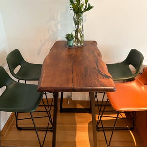 May include: A live-edge wooden table with a dark brown finish, supported by black metal legs, surrounded by four bar stools. Three stools are dark green, and one is orange. A glass vase with green plants sits on the table.