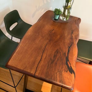 May include: A rectangular wooden table with a natural edge and dark wood grain. The table is supported by a black metal frame. Green and orange chairs surround the table. A vase of flowers and a small potted plant sit on the table.