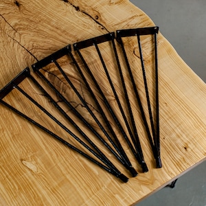 May include: A set of eight black metal hairpin legs for a table or desk. The legs are arranged in a fan shape on a light wood surface.