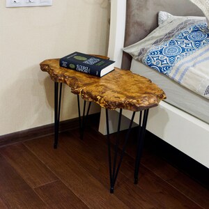 Puede incluir: Una mesa auxiliar de borde vivo con acabado de vetas de madera natural. La mesa tiene patas de horquilla negras y un libro encima. La mesa mide aproximadamente 76 cm de largo.