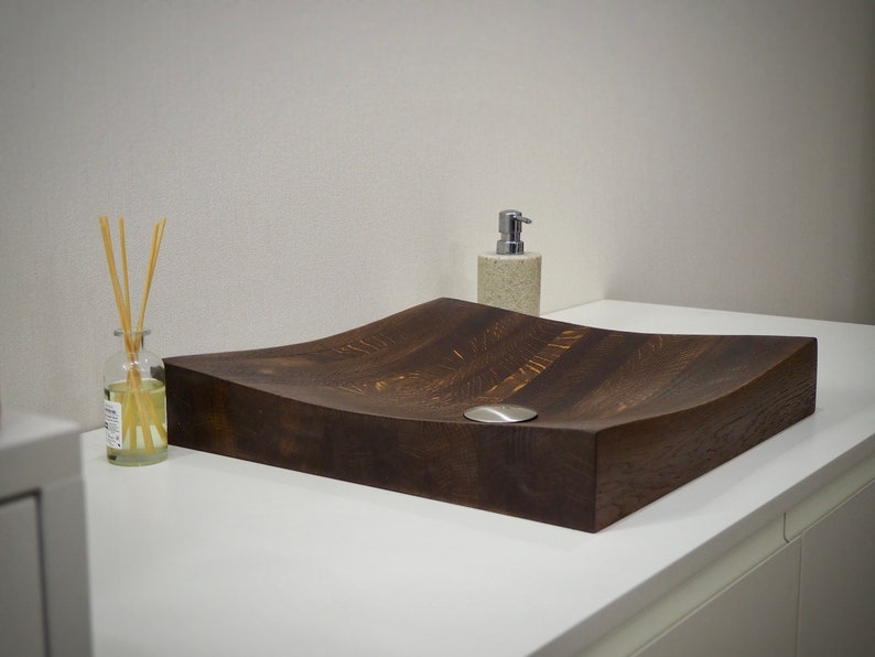 Peut inclure: Un &eacute;vier en bois brun fonc&eacute; avec un design incurv&eacute; pos&eacute; sur un comptoir blanc. L'&eacute;vier a un petit drain rond en argent.