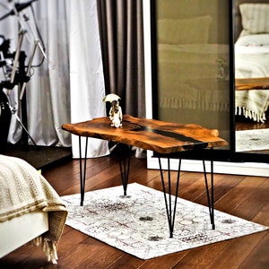 May include: A wooden coffee table with a live edge top and black epoxy resin inlay. The table has black hairpin legs and is sitting on a white and red patterned rug.