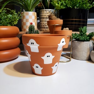 May include: A terracotta planter with a white ghost pattern. The planter is decorated with multiple white ghosts with black eyes.