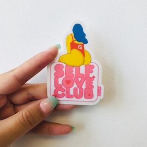 Może przedstawiać: Biała naklejka z różowym napisem "SELF LOVE CLUB" z małym sercem w środku każdej litery. Rysunek kreskówkowy osoby z niebieskimi włosami i czerwonym kostiumem kąpielowym siedzi na naklejce.