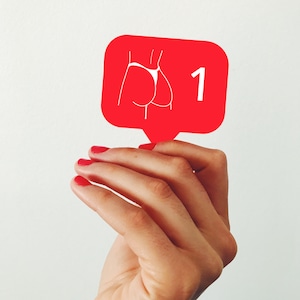 Può includere: Icona a fumetto rossa con un contorno bianco di un fondoschiena con perizoma e il numero 1. L'icona è tenuta da una mano con smalto rosso su sfondo bianco.