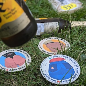 Könnte beinhalten: Drei runde Untersetzer mit Cartoon-Bildern von Hintern und Jeans, mit dem Text "I hope your day is as nice" und "Onlyakn" oder "Calubyge". Die Untersetzer liegen auf grünem Gras, mit einer Bierflasche im Hintergrund.