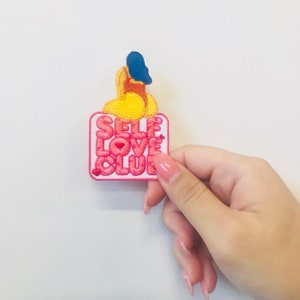 Peut inclure: Un patch brodé rose et blanc avec le texte "SELF LOVE CLUB" en rose et un petit cœur rouge. Un personnage de dessin animé jaune et bleu est au-dessus du texte.