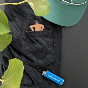 Peut inclure: T-shirt noir avec une poche, avec une figurine en bois dans la poche. Une casquette verte est dans le coin supérieur droit. Un briquet bleu avec le mot "MORNING" est en bas.