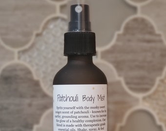 May include: A black glass bottle with a black spray nozzle and a white label that reads "Patchouli Body Mist". The label has a floral design and text that describes the product and its benefits. The bottle is sitting on a white surface.