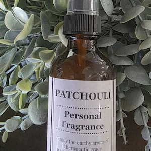 Puede incluir: Una botella de vidrio marrón con un pulverizador negro, etiquetada como "PATCHOULI Personal Fragrance". La etiqueta incluye texto sobre el aroma terroso del aceite esencial de pachulí. La botella está sobre un fondo de follaje verde.
