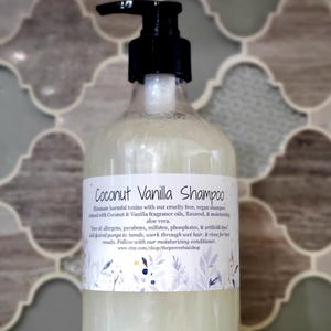Op de afbeelding: Een doorzichtige fles Coconut Vanilla Shampoo met een zwarte pompdosering. Het etiket bevat informatie over de ingrediënten en voordelen van het product. De achtergrond is een decoratief tegelpatroon.