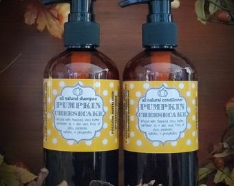 May include: Two brown bottles of shampoo and conditioner with black pumps. The labels read "Pumpkin Cheesecake" and list ingredients. The bottles are set against a backdrop of fall foliage and pinecones.