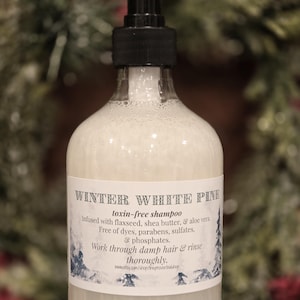 May include: A clear glass bottle of "Winter White Pine" toxin-free shampoo with a black pump. The label details ingredients such as flaxseed and shea butter. The background is blurred with green and red hues.