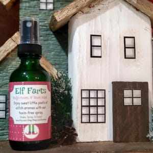 May include: A green glass bottle with a pink and white label that says "Elf Farts" and "body room, & linen mist". The label also has a cartoon elf wearing red shoes and green pants. The bottle is sitting in front of a miniature white house with brown trim.