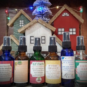 Pode incluir: Uma coleção de frascos de spray de vidro com tampas pretas, cada um rotulado com nomes festivos como "Winter White Pine" e "Candy Cane". Os frascos estão dispostos em frente a casas de madeira em miniatura e uma pequena árvore de Natal, criando um tema de feriado.