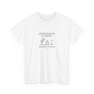 以下が含まれることがあります： 黒いグラフィックデザインの白いTシャツ。デザインは「GOWANUS CANAL SWIM TEAM」のテキストと、生き物と建物のイラストが特徴です。シャツは柔らかい素材で作られています。