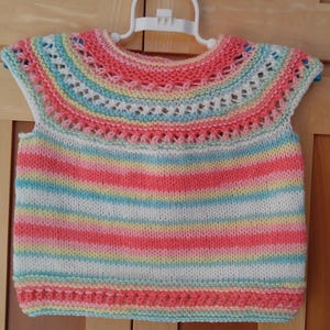 May include: A hand-knitted baby vest in a rainbow of colors, including pink, white, yellow, and blue. The vest features a round neckline with a decorative openwork pattern and is displayed on a white hanger. The striped pattern creates a cheerful and cozy look.