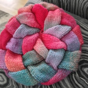 May include: A round, knitted pillow in a braided design. The pillow features a vibrant color palette of pinks, purples, and blues. The pillow is resting on a patterned gray and white surface.