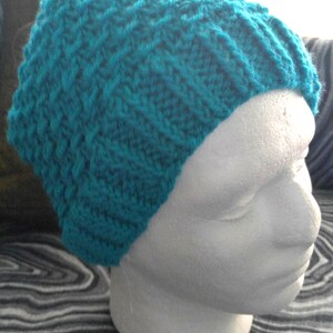May include: A teal-colored knitted beanie hat with a textured pattern. The hat is displayed on a white mannequin head, showcasing the hat's shape and design. The hat is made of a soft, textured yarn.