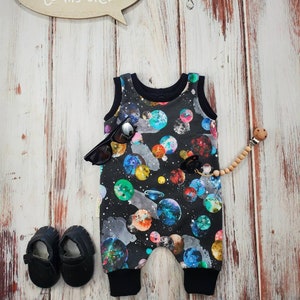 May include: A black baby romper with a colorful galaxy print, black sunglasses, and black baby shoes are displayed on a wooden surface. A wooden speech bubble sign says "WELCOME to my CRIB". A pacifier with a wooden bead is also present.