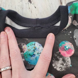 May include: A black crew neck shirt with a space-themed pattern featuring colorful planets and stars. The shirt has a black collar and a white inner lining. The planets are in shades of blue, green, pink, and gray, with speckled details.