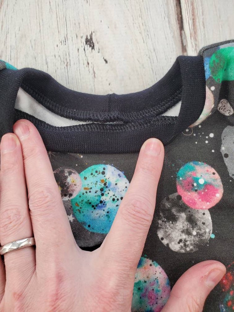May include: A black crew neck shirt with a space-themed pattern featuring colorful planets and stars. The shirt has a black collar and a white inner lining. The planets are in shades of blue, green, pink, and gray, with speckled details.