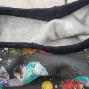 May include: Close-up of a black t-shirt with a vibrant space-themed print. The design features colorful planets, stars, and a dark blue neckline. The inner lining is white with small, scattered white dots.