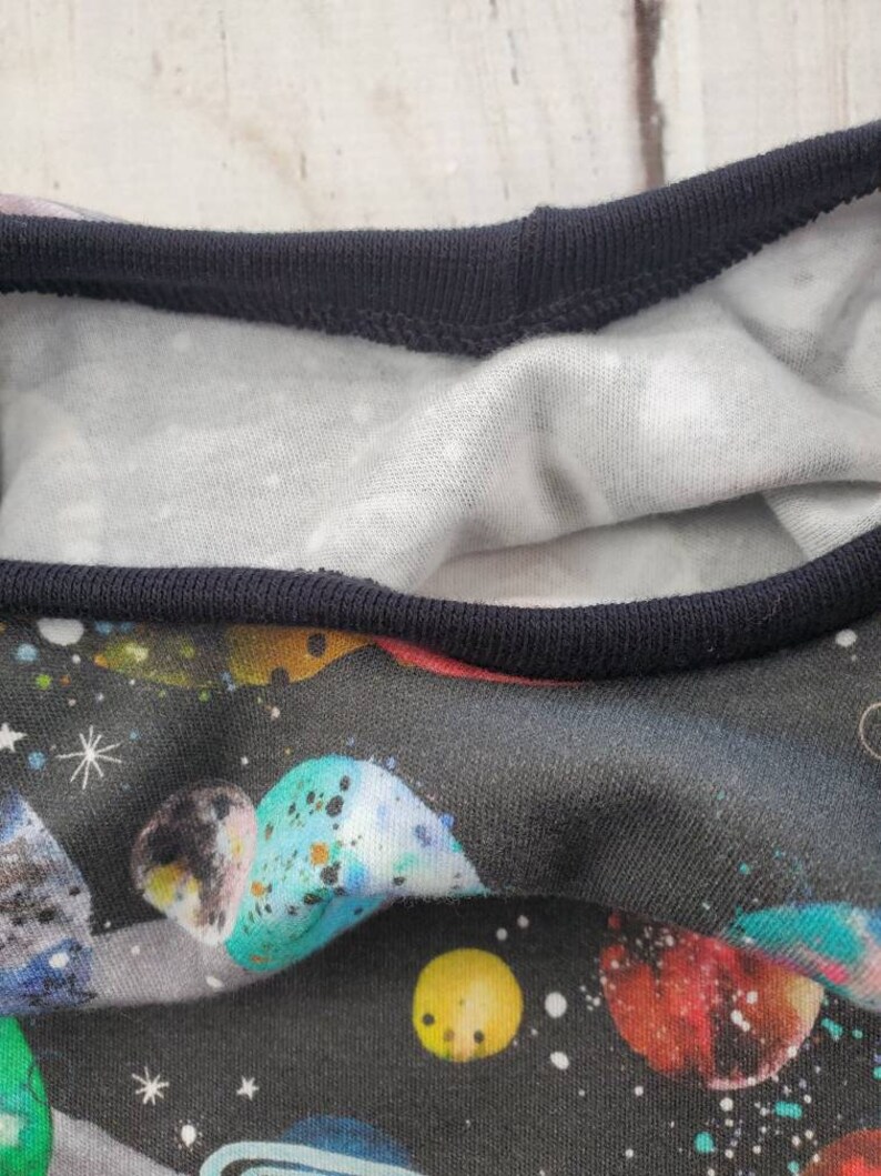 May include: Close-up of a black t-shirt with a vibrant space-themed print. The design features colorful planets, stars, and a dark blue neckline. The inner lining is white with small, scattered white dots.