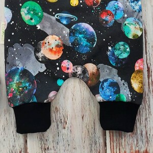 May include: Black fabric with a colorful planet pattern. The design features planets in shades of blue, green, orange, and yellow against a dark background with white stars. The cuffs are solid black.