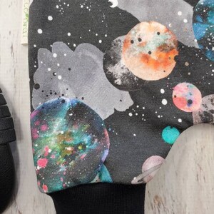 May include: A black fabric with a colorful galaxy and planet design. The planets are in shades of blue, green, orange, and pink, set against a dark background with white stars. The cuff is solid black.