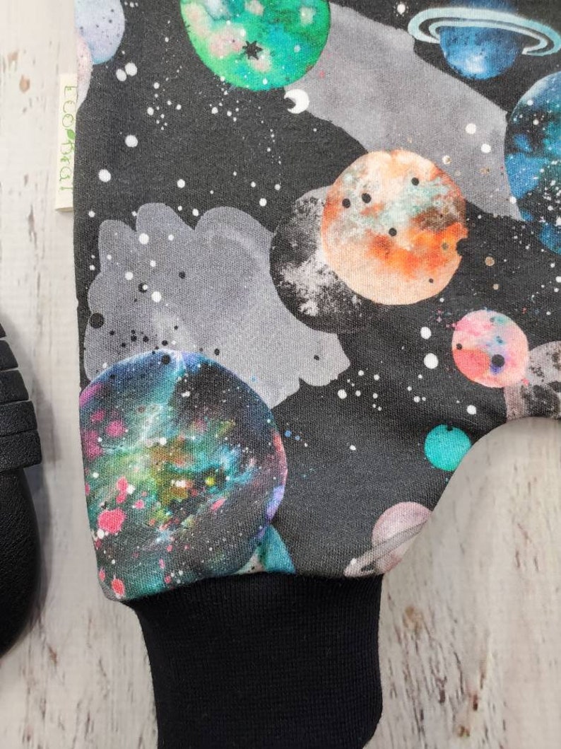 May include: A black fabric with a colorful galaxy and planet design. The planets are in shades of blue, green, orange, and pink, set against a dark background with white stars. The cuff is solid black.