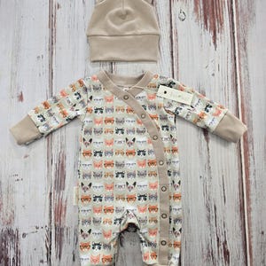 May include: A beige baby onesie with a print of cats wearing glasses. The onesie has a snap closure and fold-over cuffs. A matching beige cat-eared hat is included.