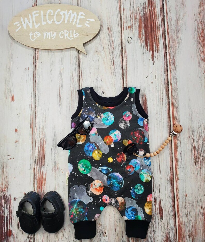May include: A black baby romper with a colorful galaxy print, black sunglasses, and black baby shoes are displayed on a wooden surface. A wooden speech bubble sign says "WELCOME to my CRIB". A pacifier with a wooden bead is also present.