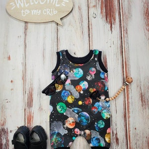May include: A black baby romper with a colorful galaxy print, black sunglasses, and black baby shoes are displayed on a wooden surface. A wooden speech bubble sign says "WELCOME to my CRIB". A wooden pacifier clip is also present.