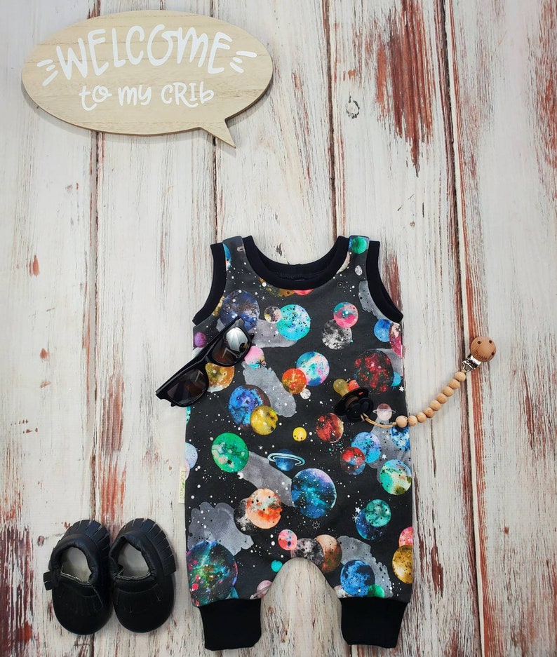 May include: A black baby romper with a colorful galaxy print, black sunglasses, and black baby shoes are displayed on a wooden surface. A wooden speech bubble sign says "WELCOME to my CRIB". A wooden pacifier clip is also present.
