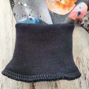 May include: Close-up of a dark blue ribbed cuff on a garment. The fabric above the cuff features a colorful space-themed print with planets, stars, and nebulas in shades of gray, orange, blue, and pink. The cuff is neatly stitched.