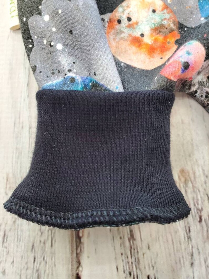 May include: Close-up of a dark blue ribbed cuff on a garment. The fabric above the cuff features a colorful space-themed print with planets, stars, and nebulas in shades of gray, orange, blue, and pink. The cuff is neatly stitched.