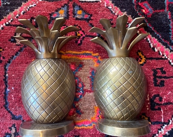 May include: Two brass pineapple sculptures with textured bodies and leafy tops. Each pineapple sits on a circular base. The sculptures are displayed on a patterned red and black rug.