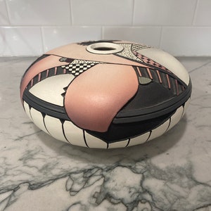 May include: A decorative, oval-shaped ceramic container with a lid. The container features a color palette of white, black, and pink, with geometric and abstract designs. The lid has a small circular opening. The container is placed on a marble surface.