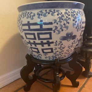 May include: A large, round, white and blue ceramic planter with a decorative floral and geometric pattern. The planter sits on a dark brown wooden stand with curved legs. The planter has a double happiness symbol.