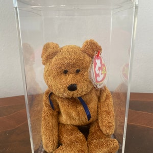 May include: A brown teddy bear with a blue ribbon around its neck, displayed in a clear plastic case. The bear has a black nose and a red and white tag attached. The plush toy is a classic collectible.