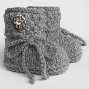May include: A pair of gray knitted baby booties. Each bootie features a decorative silver button and a knitted tie. The booties are made of a soft, textured yarn, perfect for keeping little feet warm.