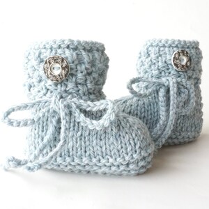 May include: A pair of light blue knitted baby booties. Each bootie features a decorative silver button and a matching knitted tie. The booties are made of soft yarn and are designed for newborns.
