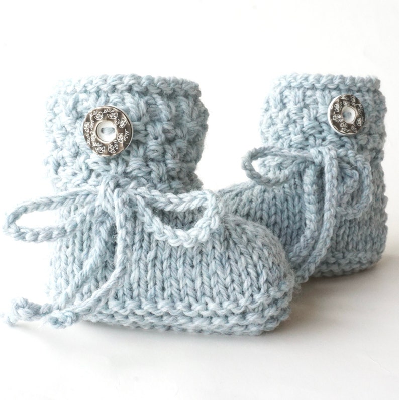 May include: A pair of light blue knitted baby booties. Each bootie features a decorative silver button and a matching knitted tie. The booties are made of soft yarn and are designed for newborns.