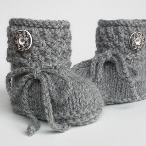 May include: A pair of gray knitted baby booties. Each bootie features a decorative silver button and a knitted tie. The booties are made of a soft, textured yarn, perfect for keeping little feet warm.