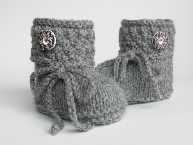 May include: A pair of gray knitted baby booties. Each bootie features a decorative silver button and a knitted tie. The booties are made of a soft, textured yarn, perfect for keeping little feet warm.