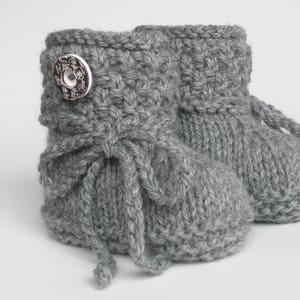 May include: A pair of gray knitted baby booties. Each bootie has a decorative button and a tie closure. The booties are made of a soft, textured yarn, perfect for keeping little feet warm. The overall design is simple and classic.