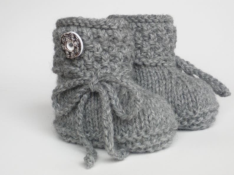 May include: A pair of gray knitted baby booties. Each bootie has a decorative button and a tie closure. The booties are made of a soft, textured yarn, perfect for keeping little feet warm. The overall design is simple and classic.