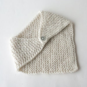 Può includere: Una sciarpa di maglia color crema con chiusura a bottone. La sciarpa è piegata a metà e il bottone è visibile al centro.